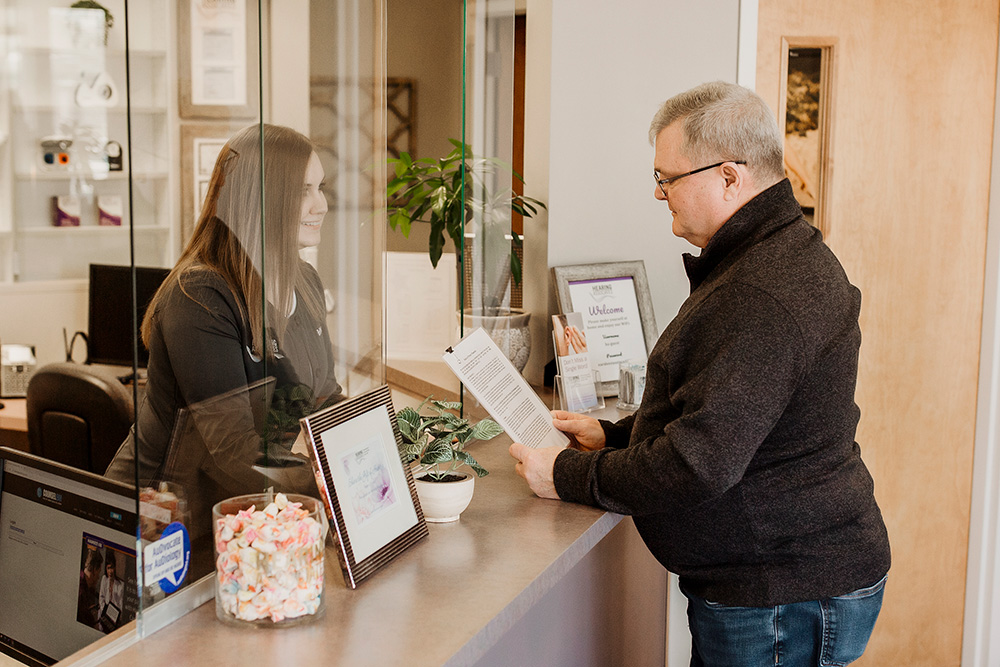 A man stands at a reception desk reading a document while a receptionist smiles at him from behind a glass barrier. There are framed pictures, a potted plant, and a jar of candies on the counter.