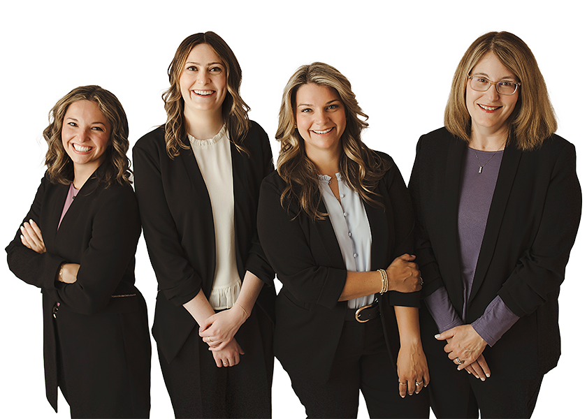 Four women standing side by side, smiling, and dressed in business attire with black blazers against a white background.