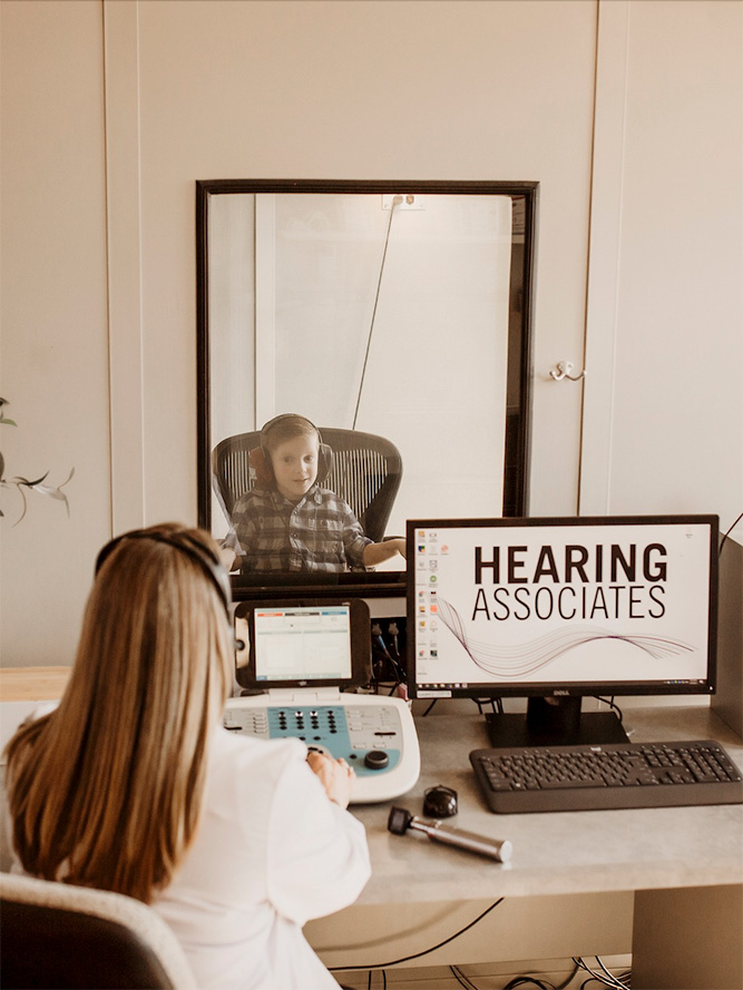 A young boy sits in a sound booth facing a woman at a desk with hearing test equipment. A computer monitor reads Hearing Associates. They are separated by a glass window.