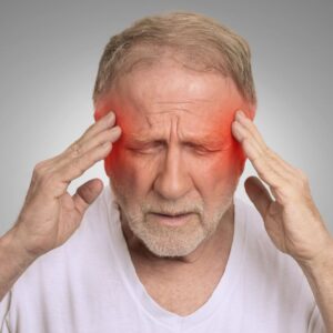 Older man with gray hair holds his temples and closes his eyes in pain, with red coloring on both sides of his head indicating a headache or migraine. He wears a white shirt and appears distressed.